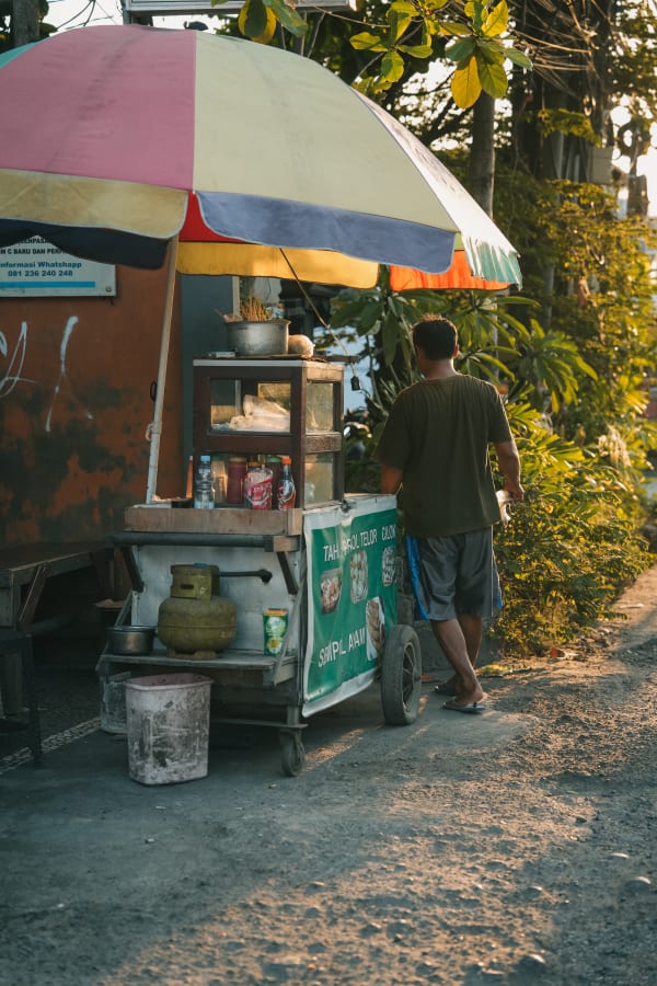 Bali street food vendor next to colorful cart with golden light, selling Tahu Telor, Cilok, Sempol Ayam. Denpasar, Indonesia.