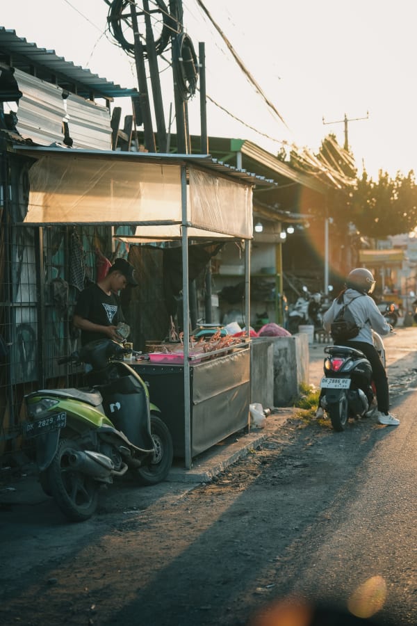 Bali street scene at golden hour with local vendor at market stall selling poultry, two scooters parked on a dusty roadside in Indonesia.