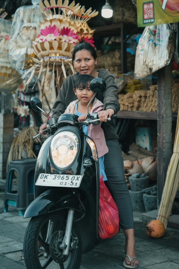 Balinese woman and young child on black scooter, surrounded by vibrant palm leaf offerings in a lively market street, Bali, Indonesia.