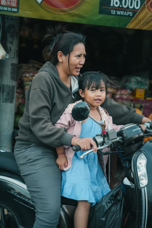 Candid street portrait of a Balinese woman and young girl on a black Honda scooter, market background, Bali, Indonesia.