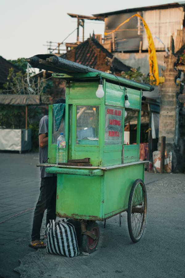 Green Sate Ayam Lontong Madura street food cart in a Bali village, Indonesia. Vendor prepares food, traditional architecture, yellow penjor.