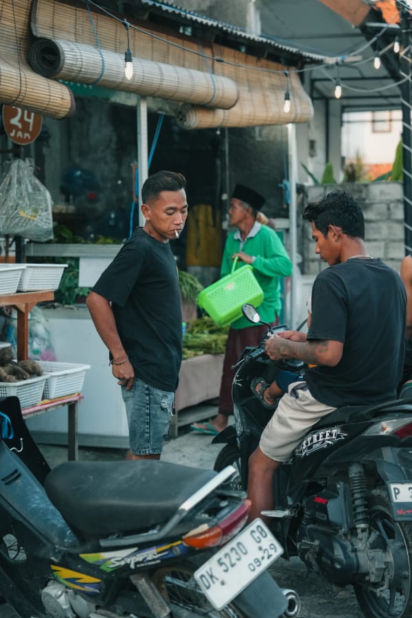 Three Balinese men, one smoking, two with scooters, near a small roadside stall with bamboo blinds and string lights in Bali, Indonesia.