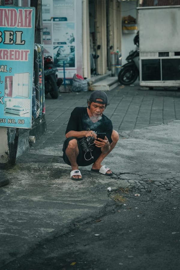 Bali street scene, local man squatting, smoking, using smartphone, urban setting, cracked concrete, faded shop sign, Indonesia.