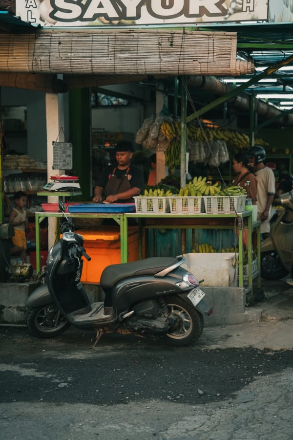 Bali street market scene with male vendor, fresh green bananas, produce stalls, and a grey Honda Scoopy scooter parked on pavement.