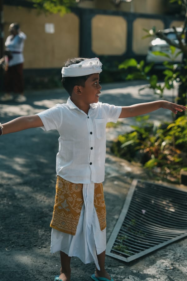 Balinese boy in traditional white shirt, patterned yellow sarong, and headwear, arms outstretched on a sunny street in Bali.