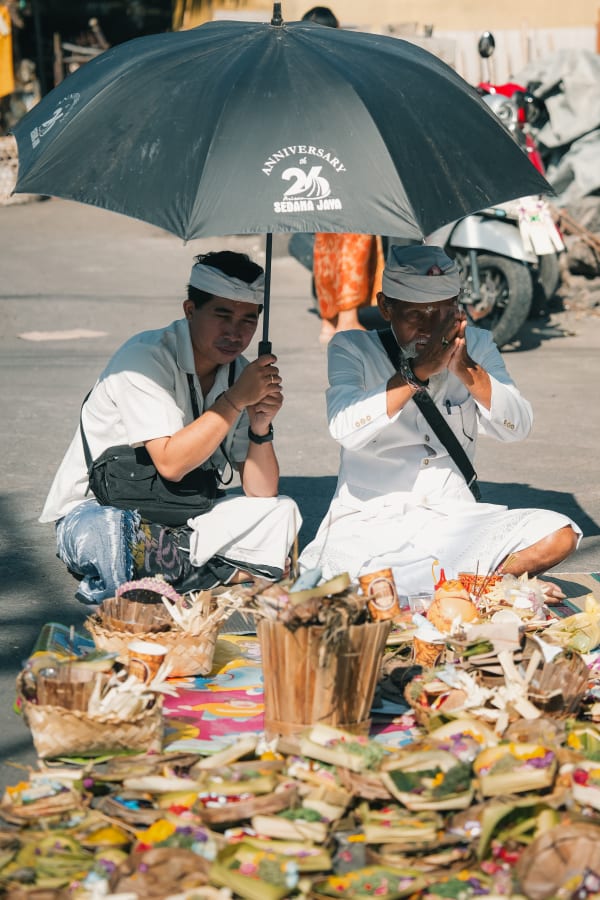 Two Balinese men in traditional attire, shaded by an umbrella, perform a street prayer with vibrant offerings in Bali, Indonesia.
