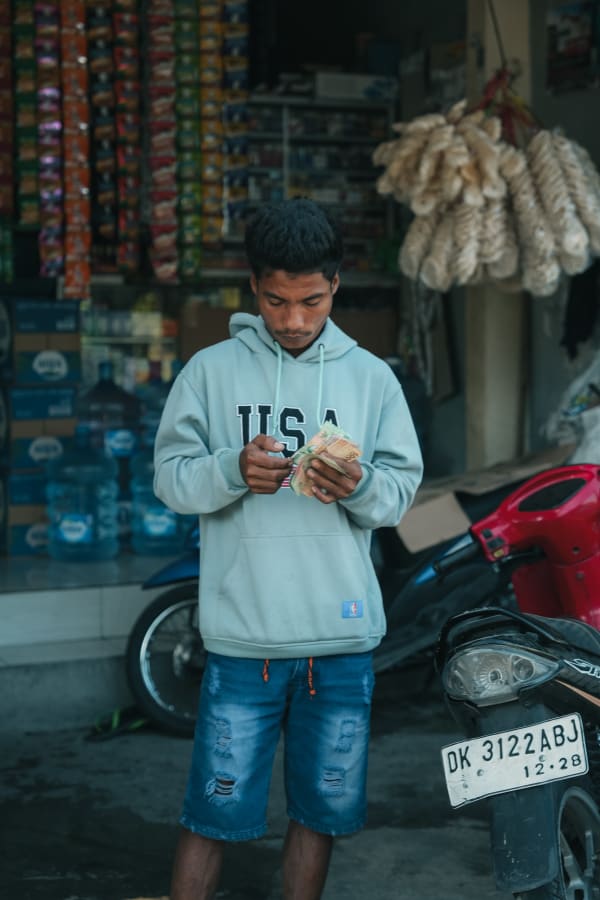 Young Indonesian man counting rupiah outside a Bali street shop with stacked snacks, water bottles, and a parked scooter displaying a 'DK' license plate.