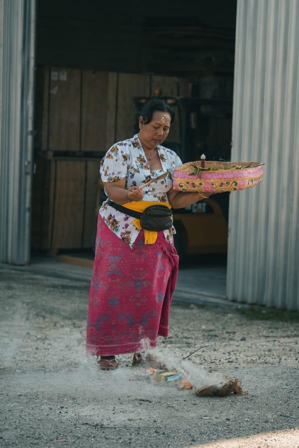Balinese woman in traditional attire performs a ceremony with smoking offerings, holding a woven basket on a gravel path in Bali, Indonesia.