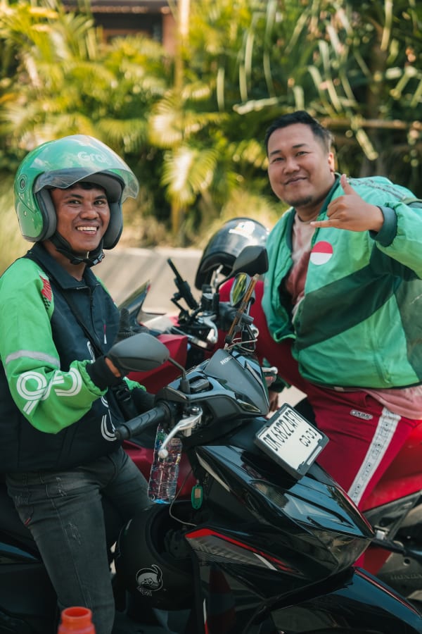 Two smiling Indonesian Grab motorcycle drivers in green uniforms, sitting on Honda scooters, tropical Bali street background.