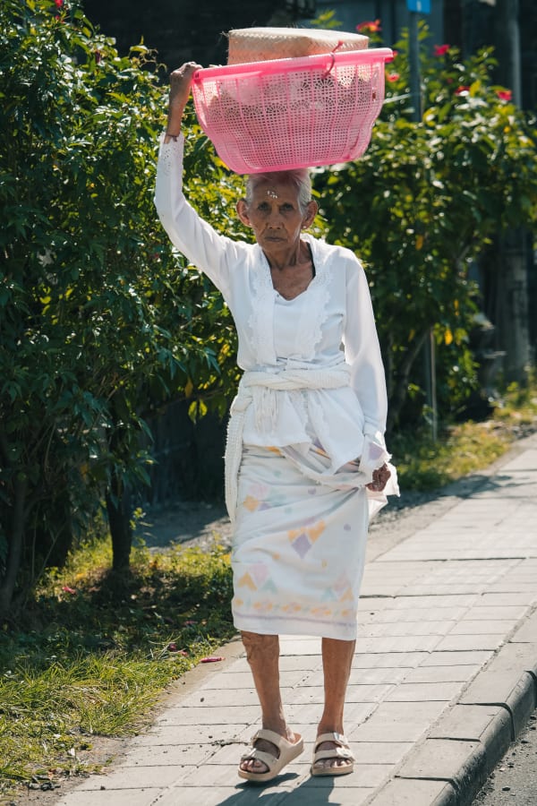 Elderly Balinese woman carrying a pink basket on her head, walking on a sunny sidewalk in Bali, Indonesia. Traditional white attire, green foliage background.