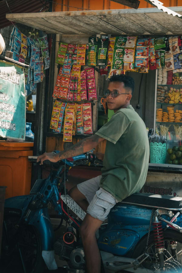 Young man on blue scooter at a vibrant Bali street warung, displaying instant drink sachets and snacks, smiling at camera.