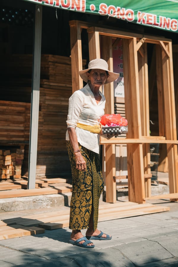 Elderly Balinese woman in traditional sarong and straw hat holding offerings, standing on a sunny street near Sono Keling wood shop in Bali.