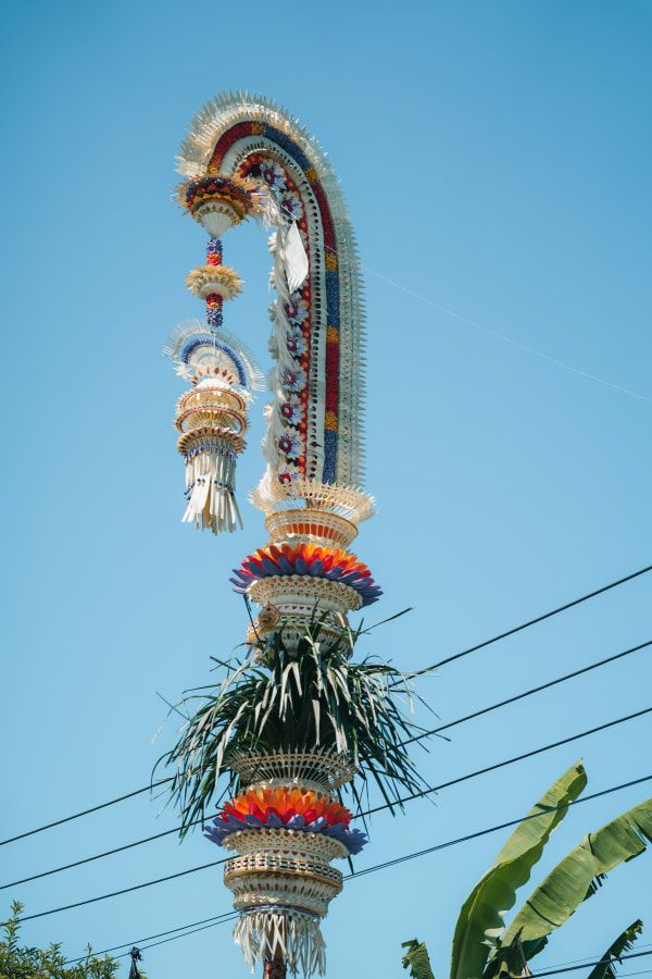 Balinese Penjor pole against clear blue sky, intricate bamboo weaving, vibrant offerings, green foliage, power lines, Bali, Indonesia