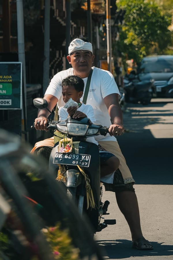 Balinese father and young son riding scooter, man in traditional udeng headwear, boy holding currency, street scene, Bali, Indonesia