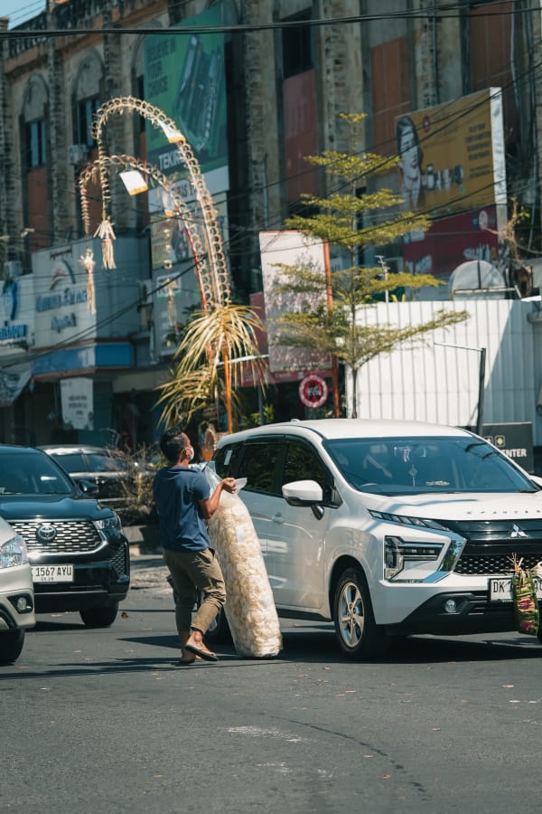 Man in mask carries large krupuk bag past white car on sunny Bali street, traditional decorations visible. Indonesia.