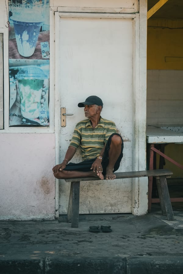 Elderly Balinese man in striped shirt, cap, sitting barefoot on wooden bench outside shop with Ultra Milk posters, Bali, Indonesia.