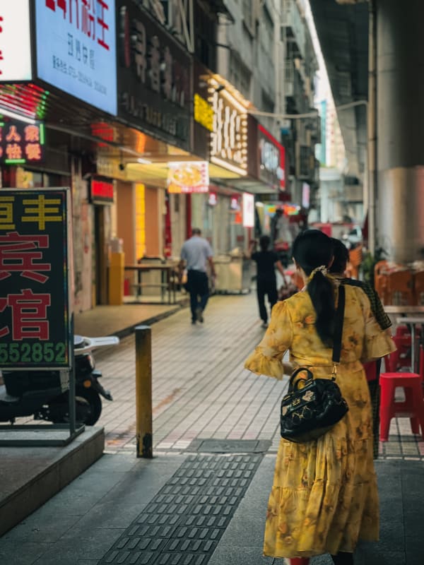 Woman in yellow floral dress walks a lively Longgang District street in China, past neon storefronts and blurred figures at dusk.