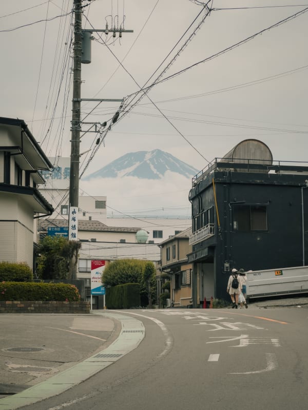 Mount Fuji, partly cloud-covered, glimpsed above a street in Fujikawaguchiko, Japan. Power lines, Japanese buildings, pedestrians.