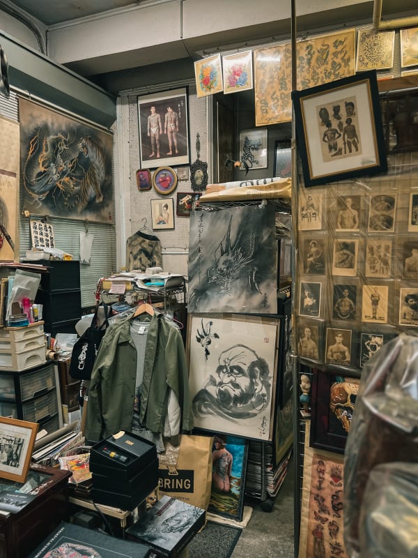 Cluttered traditional Japanese tattoo studio interior in Yokohama, Japan, featuring irezumi flash art, paintings, and vintage photography.