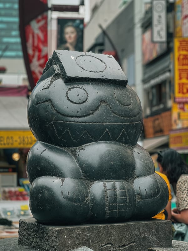 Mysterious dark stone sculpture with stylized smiling face and block hat, on a bustling Taito, Tokyo street.