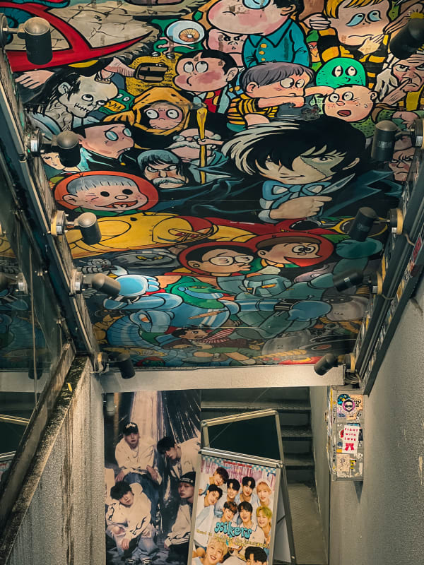 Colorful manga anime mural ceiling in Shibuya, Japan, with K-pop posters, urban art, and reflective walls.
