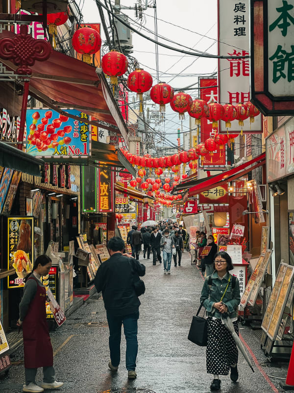Bustling Yokohama Chinatown street in Japan with vibrant red lanterns, crowded shops, and people walking on wet pavement under an overcast sky.