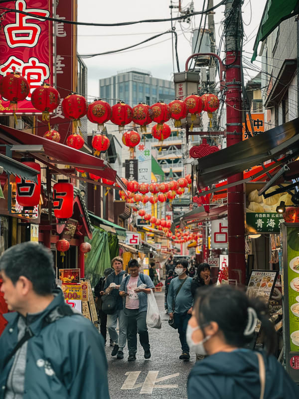 Bustling street scene in Yokohama Chinatown, Japan, filled with red lanterns, vibrant shop signs, and pedestrians under an overcast sky.