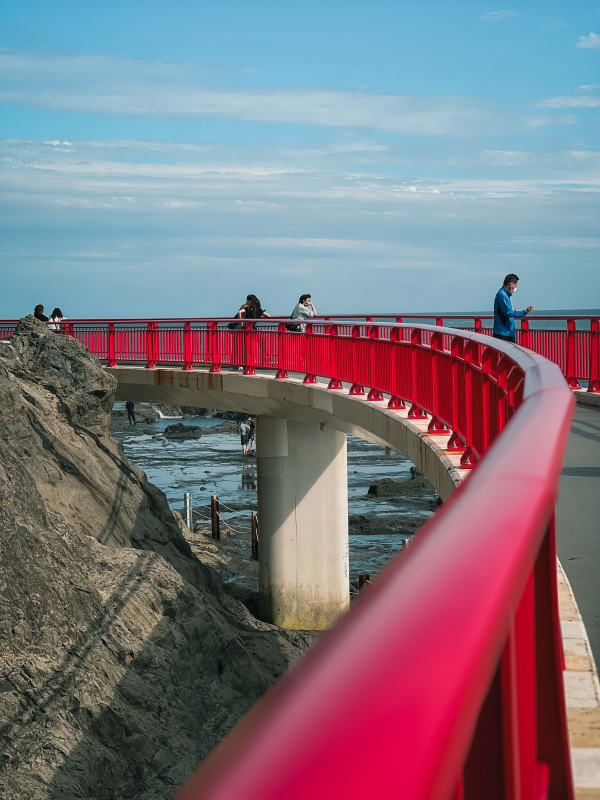 Enoshima Island red pedestrian bridge over rocky coast, Fujisawa, Japan. People enjoy ocean views under blue sky.