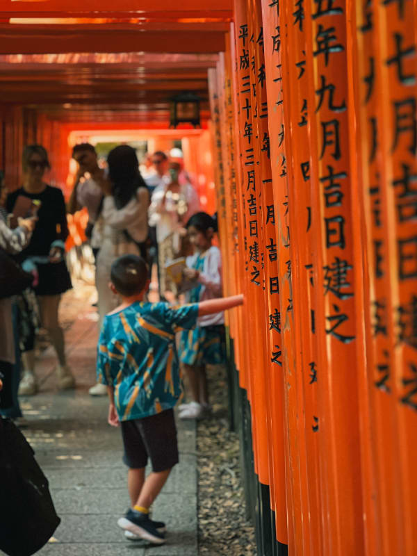 Young boy touches vibrant orange torii gate post at Fushimi Inari Taisha, Kyoto. Japanese script visible on traditional path.