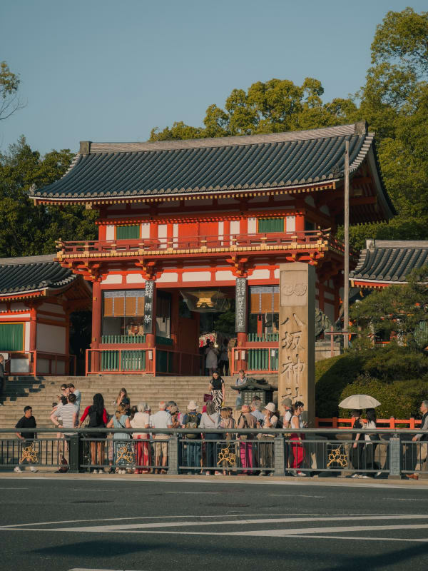 Heian Shrine Ōtenmon gate in Kyoto, Japan, with tourists gathered. Vermillion architecture, green roofs, warm afternoon light.