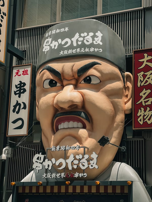 Kushikatsu Daruma giant chef head sign in Osaka, Japan. Stern face, mustache, bold Japanese kanji, vibrant street food icon.