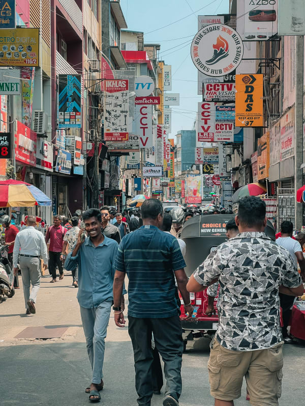 First Cross Street, Pettah, Colombo, Sri Lanka: Bustling street scene with crowded pedestrians, vibrant electronics and shoe shop signs, a man smiling on phone.
