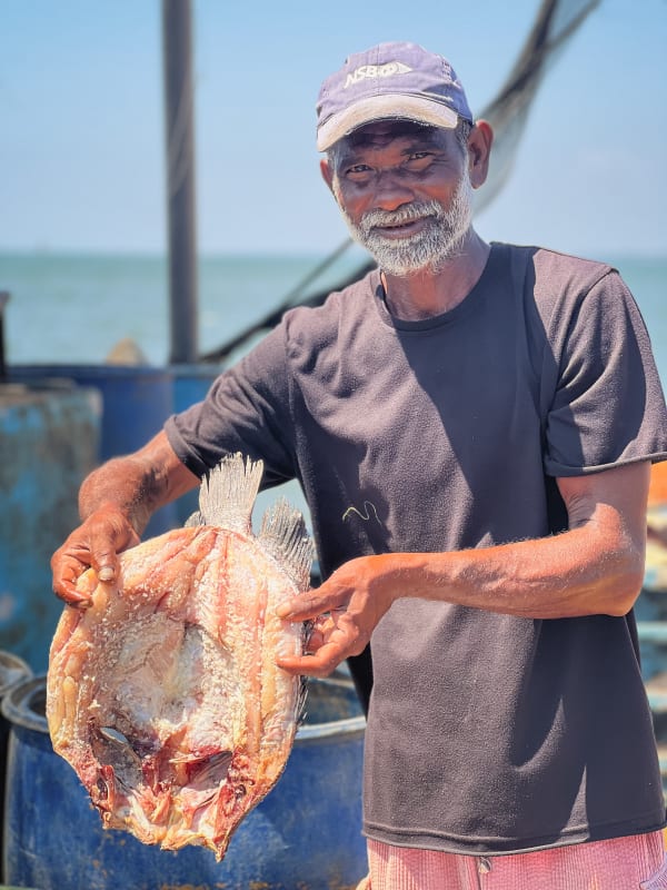 Smiling Sri Lankan fisherman in Negombo holding a large, sun-dried fish, with ocean background. Traditional seafood preparation.