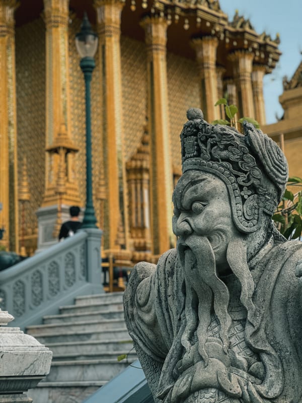 Intricate stone guardian statue, bearded, ornate golden temple columns, Grand Palace, Bangkok, Thailand.