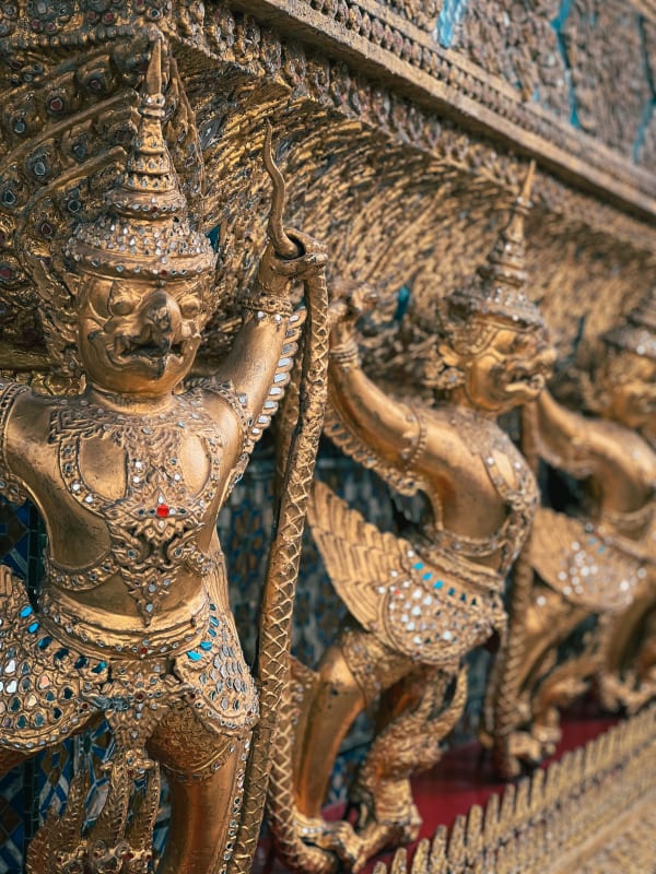 Golden Garuda statues adorned with mirrored mosaics and jewels at Grand Palace, Bangkok. Intricate Thai temple architecture.