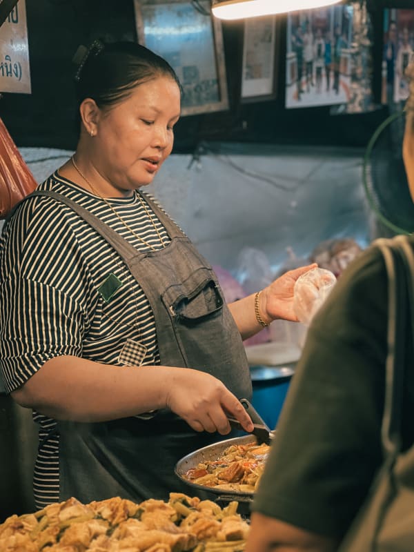 Bangkok street food vendor preparing stir-fry for a customer under warm market lights, surrounded by delicious Thai dishes.