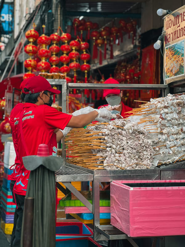 Bangkok Chinatown street food vendor prepares grilled squid skewers on ice, surrounded by red lanterns.