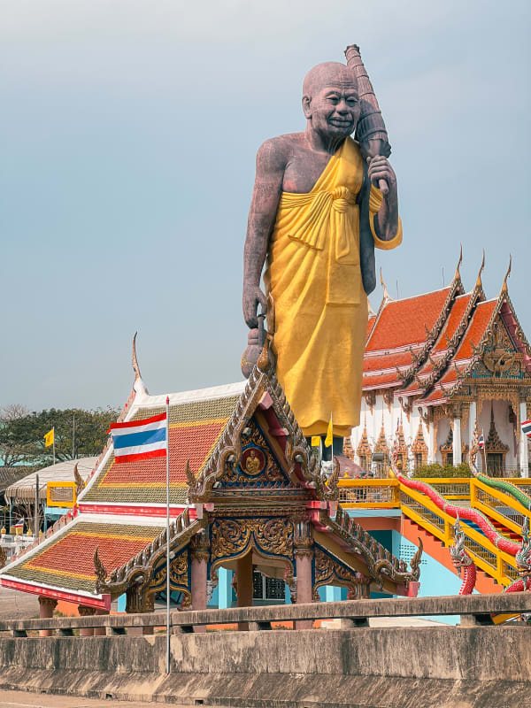 Towering Luang Pu Toh monk statue at Wat Hong Thong, Bang Pakong, Thailand, under a clear sky, vibrant temple roofs.
