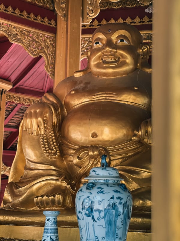 Golden laughing Buddha statue, serene expression, temple interior with red and gold ornate carvings, Bang Pu, Thailand.