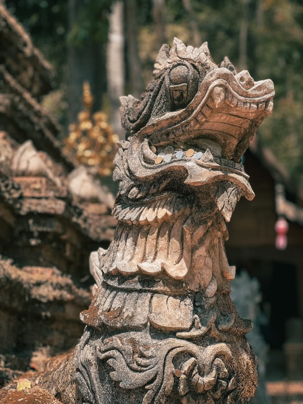 Close-up of weathered stone Naga sculpture with coins in crevices, intricate details, moss, Chiang Mai, Thailand temple art.