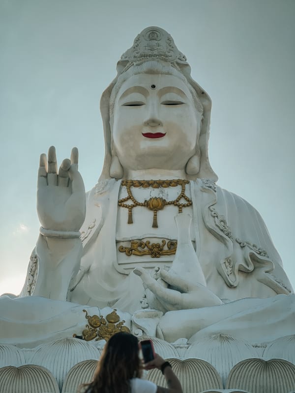 Giant white Guan Yin statue at Wat Huay Pla Kang, Chiang Rai, Thailand. Woman photographs the towering Buddhist deity against a bright sky.