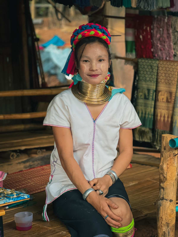 Kayan long-neck woman, Chiang Rai, Thailand. Smiling, adorned with brass neck coils, vibrant headpiece, silver bangles, white traditional dress.