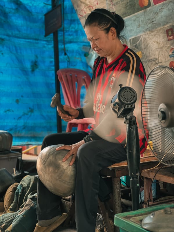 Bangkok artisan, a woman in an AC Milan jersey, hammering a metal bowl in a humble workshop. Blue tarp background, blurred fan.