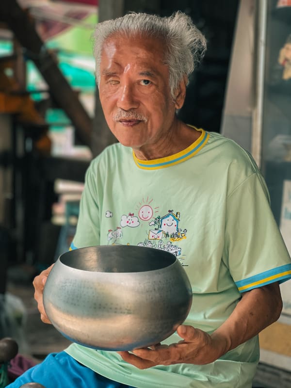 Elderly Thai man with white hair, mustache, holds a metal alms bowl in Bangkok. Vibrant street portrait, cultural scene.
