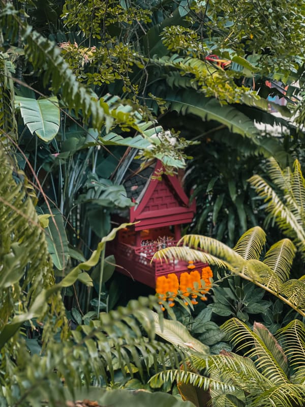 Traditional red Thai spirit house (San Phra Phum) with orange flower garlands nestled among lush tropical foliage in Bangkok, Thailand.