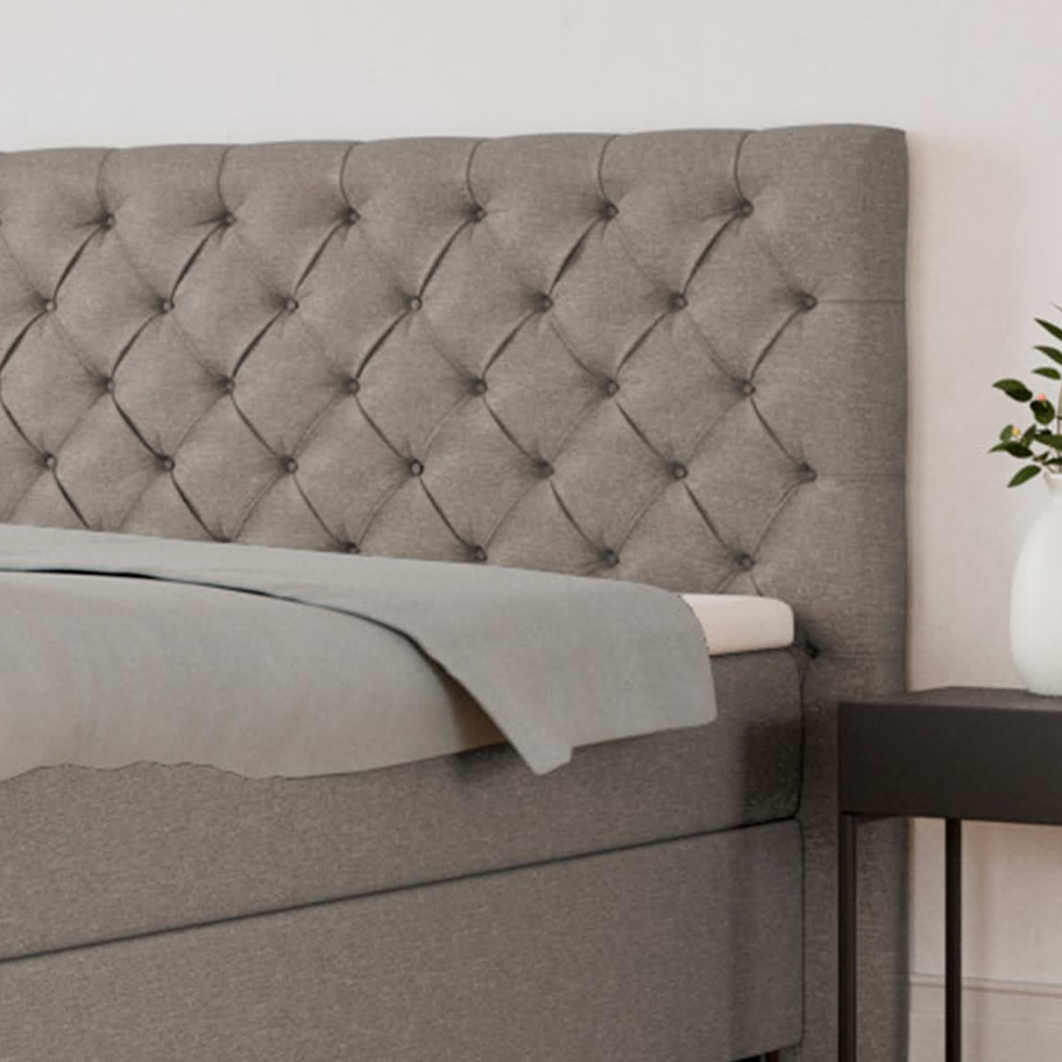 Grå polstret seng med knappet sengegavl, lysegråt sengetæppe og natbord til højre.