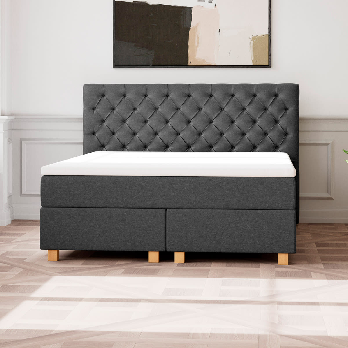 Mørkegrå kontinentalseng med polstret hovedgærde og hvid topmadras på et lyst trægulv.