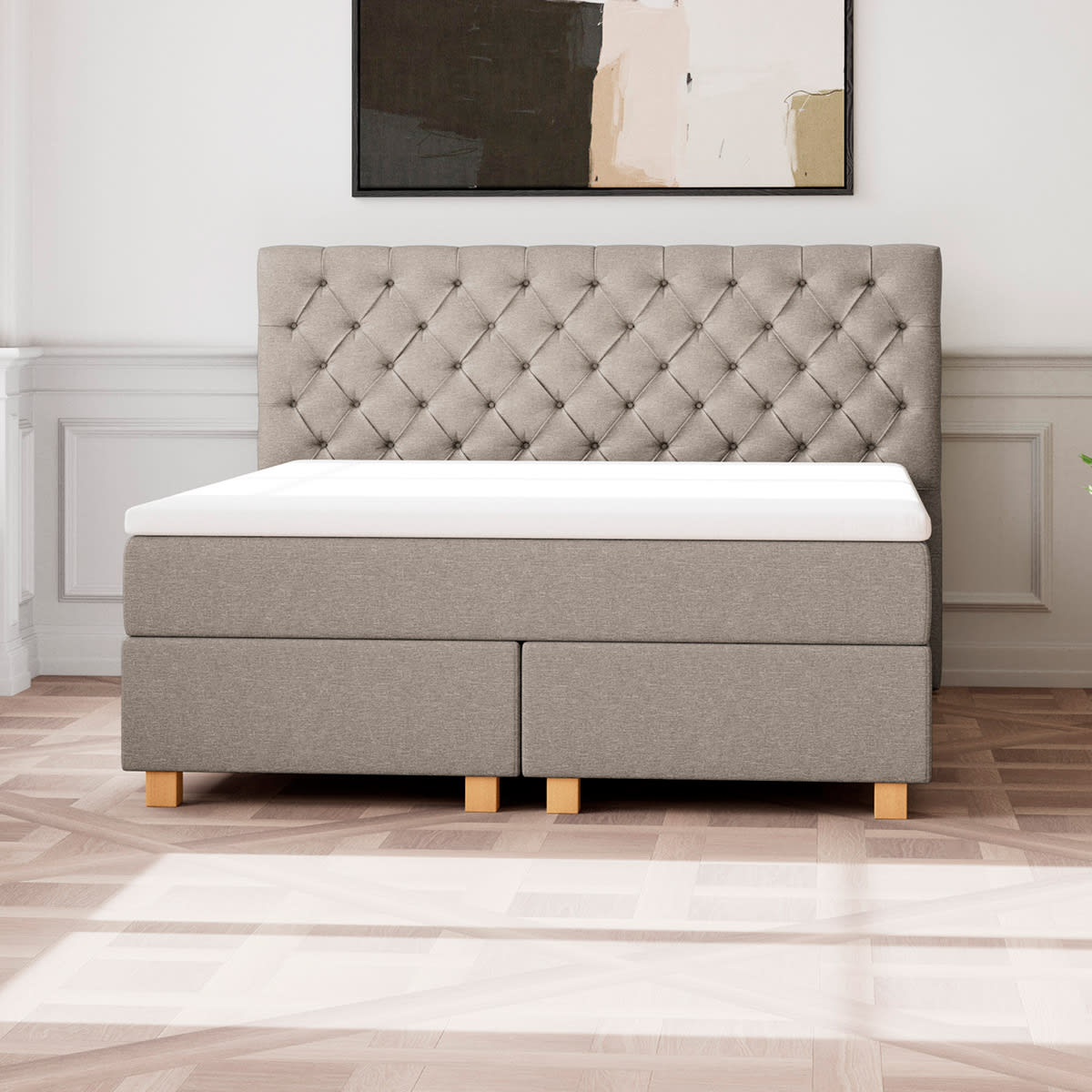 Grå kontinentalseng med polstret, tuftet sengegavl og hvid topmadras på et trægulv.