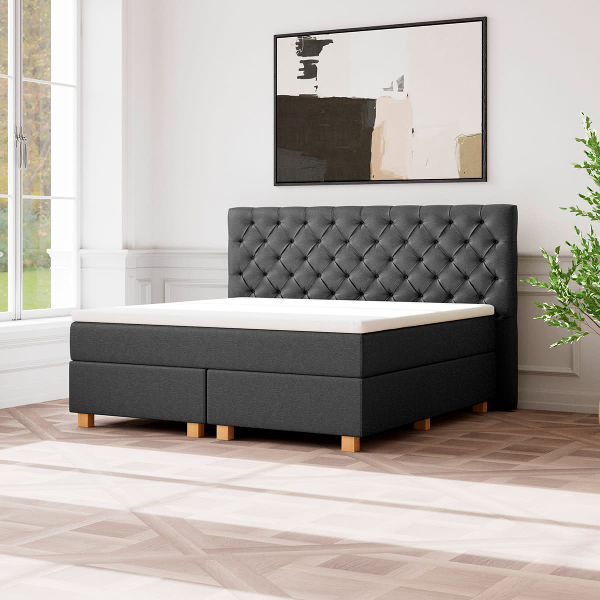 En mørkegrå kontinentalseng med polstret sengegavl, hvid topmadras og træben i et lyst rum.