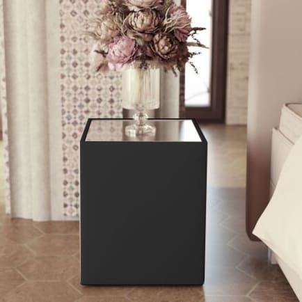 Sort kubeformet sengebord med spejlplade og en vase med lyserøde blomster.